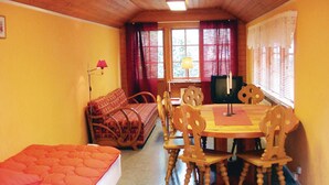Ein gemütliches Zimmer mit Holzbalkendecke, einem Bett mit orangefarbener Decke, einem Esstisch mit Stühlen, einem Fernseher und einem Fenster mit Vorhängen.