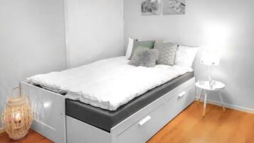 Una cama con colchón gris y ropa de cama blanca, una mesita de noche pequeña con una lámpara, y un cuadro colgado en la pared.