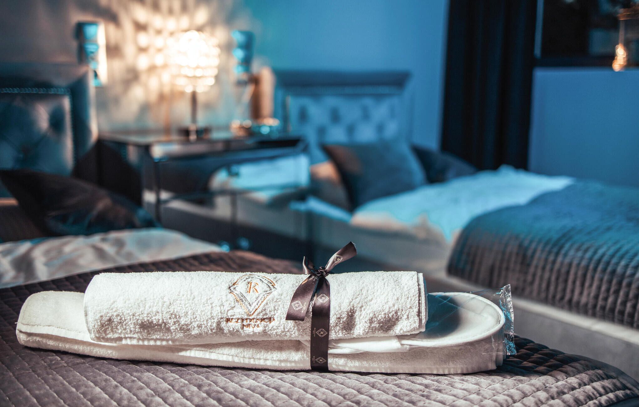 A neatly made bed with a rolled towel and a decorative ribbon, a bedside table with a lamp, and a window with curtains.