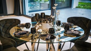 Mesa redonda de vidrio dispuesta con vajilla negra y plateada, incluyendo platos, tazones y tazas, rodeada de sillas tapizadas oscuras.