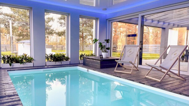 Una piscina interior moderna con paredes de vidrio transparente, un deck de madera y sillones de descanso.