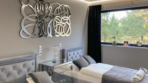 Un dormitorio moderno con un cabecero capitonado, una mesita de noche de vidrio, un adorno de pared y una ventana con vistas a árboles.