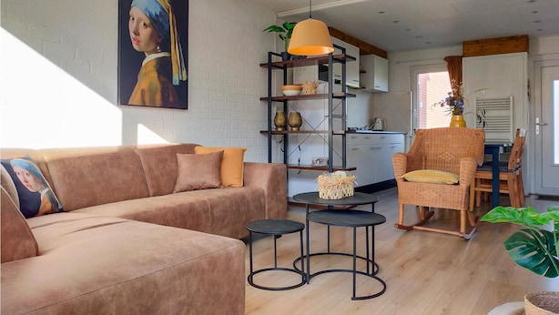 A living room with a sofa, two side tables, a wicker chair, a hanging pendant light, and a kitchen in the background.