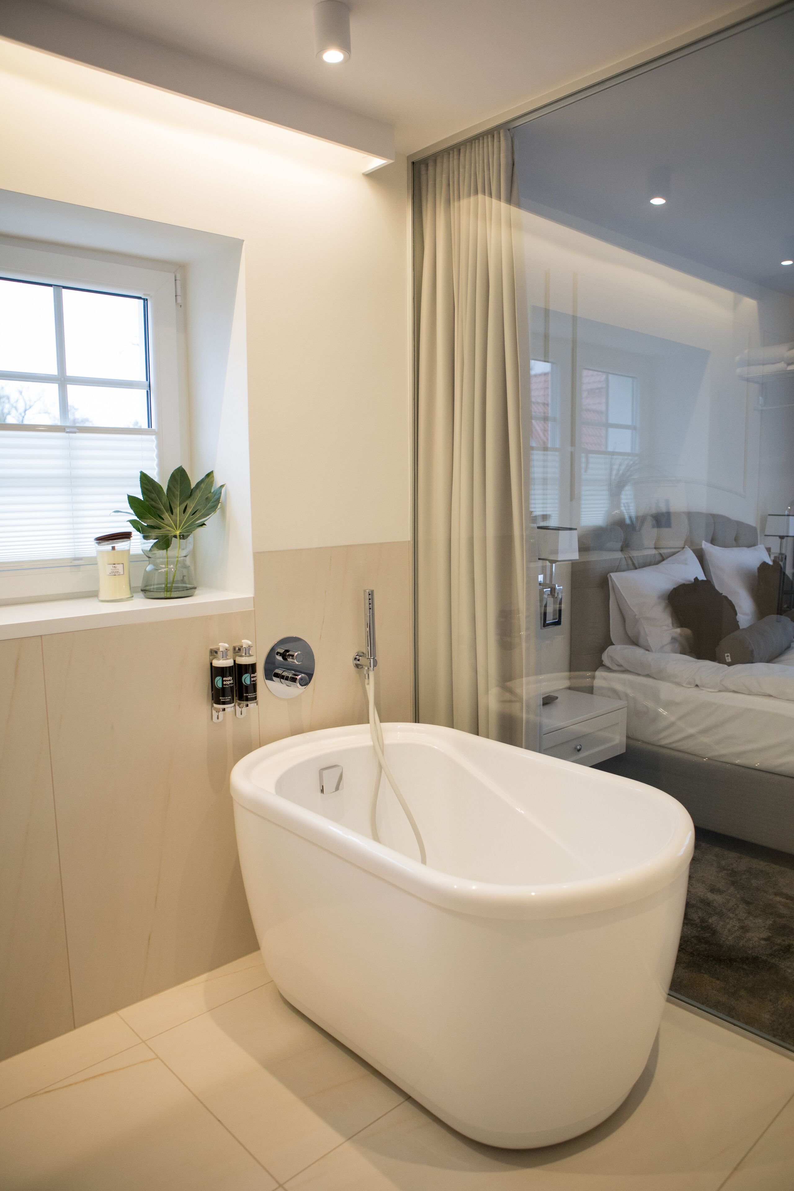 Ein modernes Badezimmer mit freistehender Badewanne, einem Fenster mit Jalousien, einer Topfpflanze und einem Nachttisch mit Lampe.