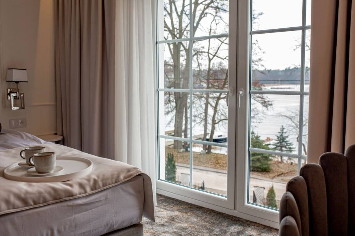 A hotel room with a large window, a bed with a tray of cups, a chair, and a lamp on the wall.
