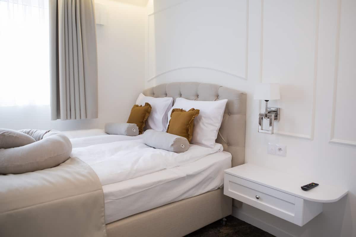 A neatly made bed with white and brown pillows, a bedside table with a remote control, and a wall-mounted lamp.