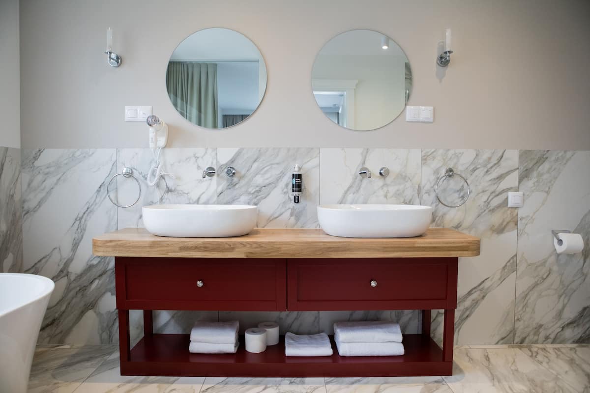 A bathroom with two sinks, marble backsplash, wooden vanity, and a red cabinet below.