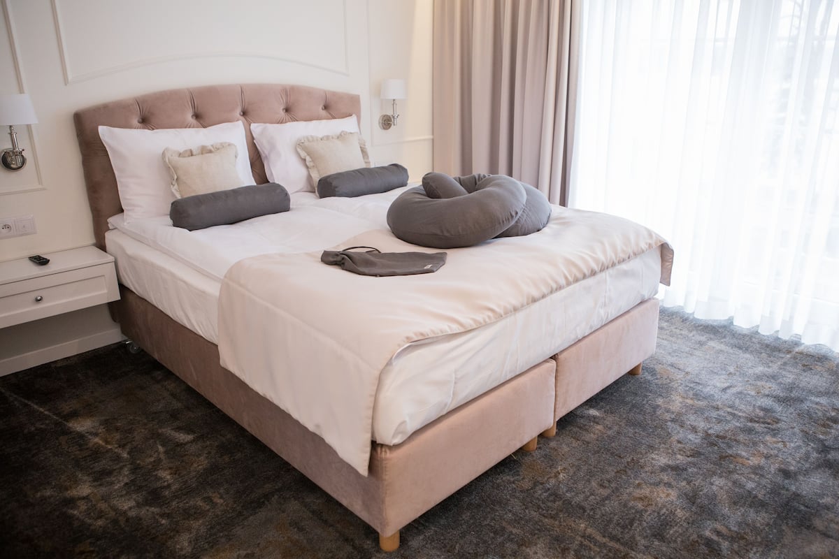 A neatly made bed with a tufted headboard, a bedside table with a lamp, and a large window with sheer curtains.