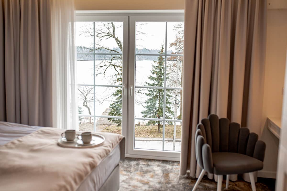 A hotel room with a bed, a chair, a window with a view of trees and mountains, and a tray with cups on the bed.