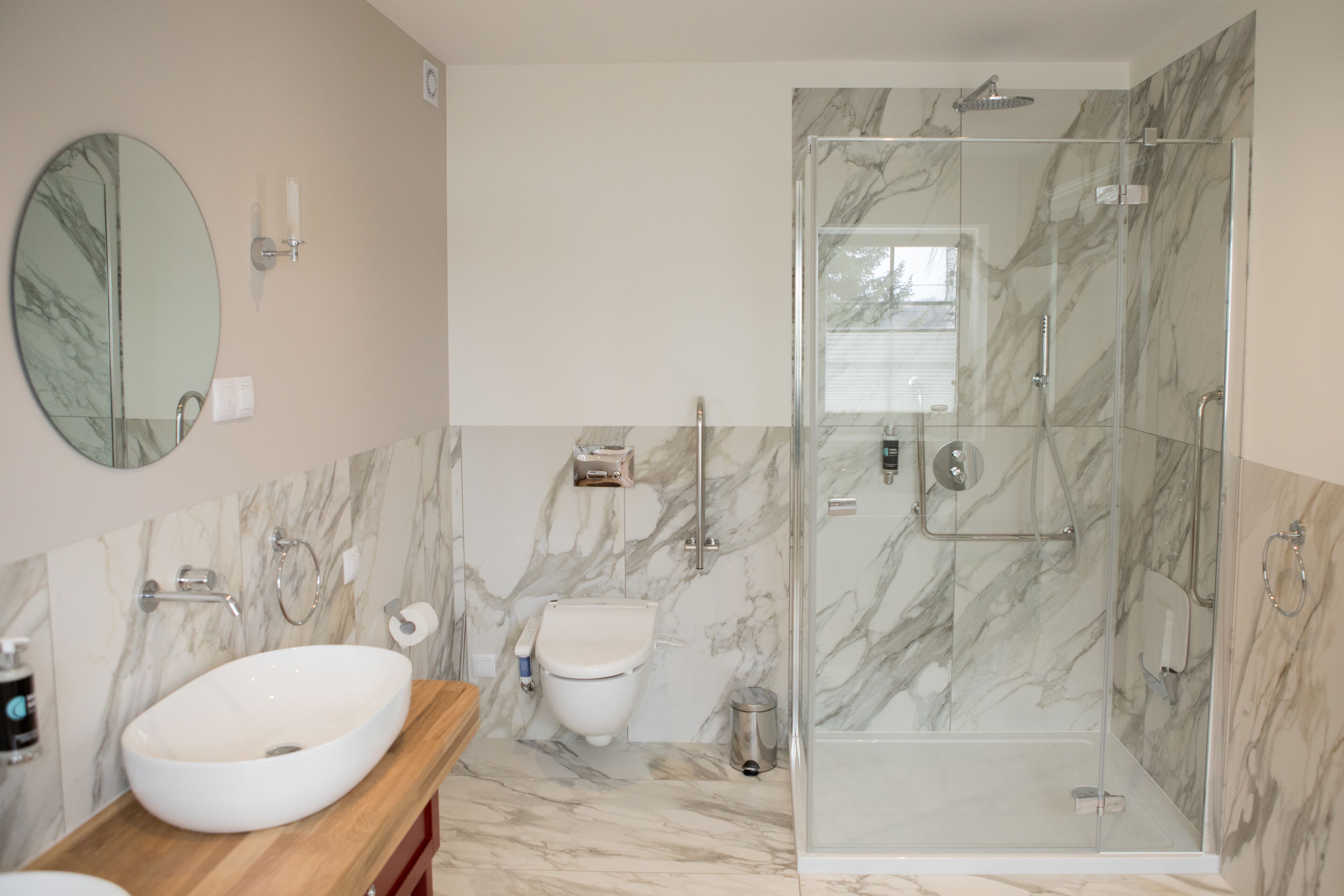 Un baño moderno con suelo de mármol, un lavamanos ovalado grande, un inodoro y una ducha con mampara de vidrio.