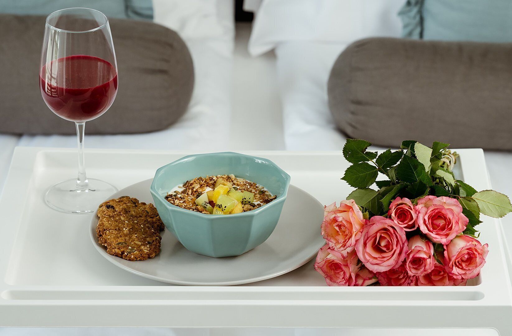 Ein Tablett mit einem Glas Rotwein, einer Schüssel Granola mit Obst, einem Keks und einem Strauß rosa Rosen.
