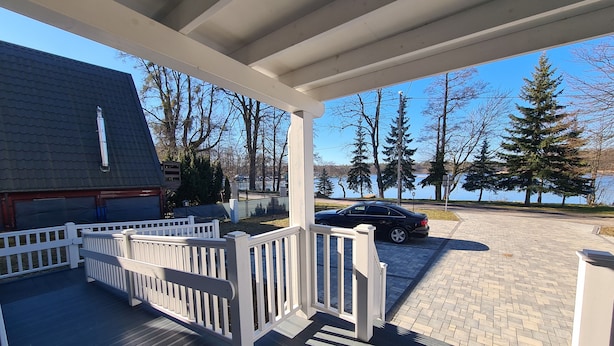 A covered porch with a white railing, a paved driveway, and a view of a lake and trees. - Maly Sopot Boutique (Ilawa)