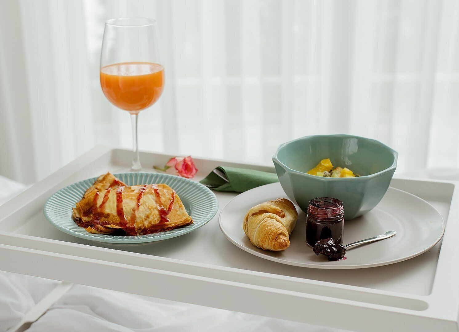 A tray with a pastry, a croissant, a bowl of fruit, a glass of orange juice, and a jar of jam.