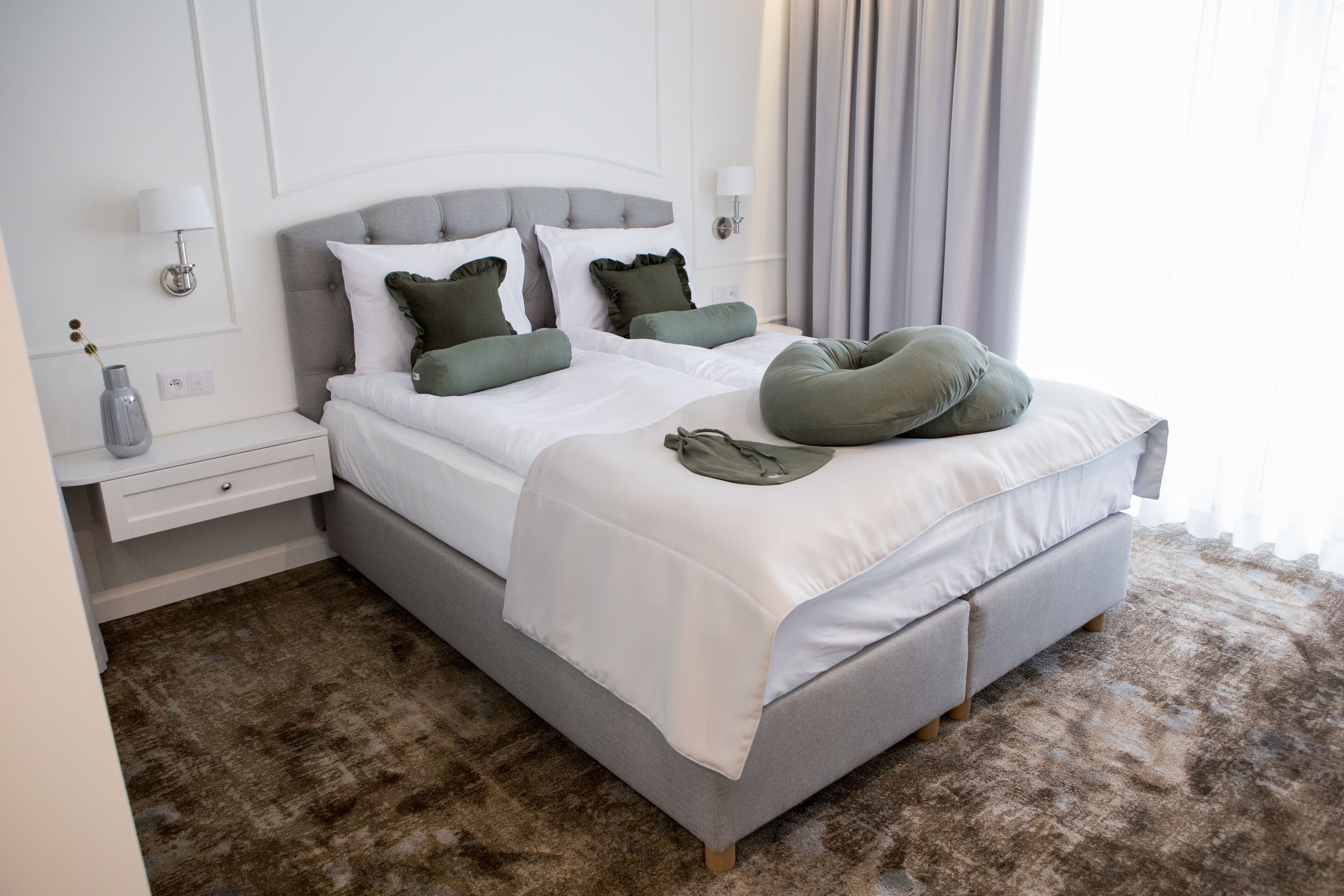 A neatly made bed with a grey headboard, white bedding, and green pillows. There is a bedside table with a lamp and a vase. The room has a brown carpet and a window with curtains.