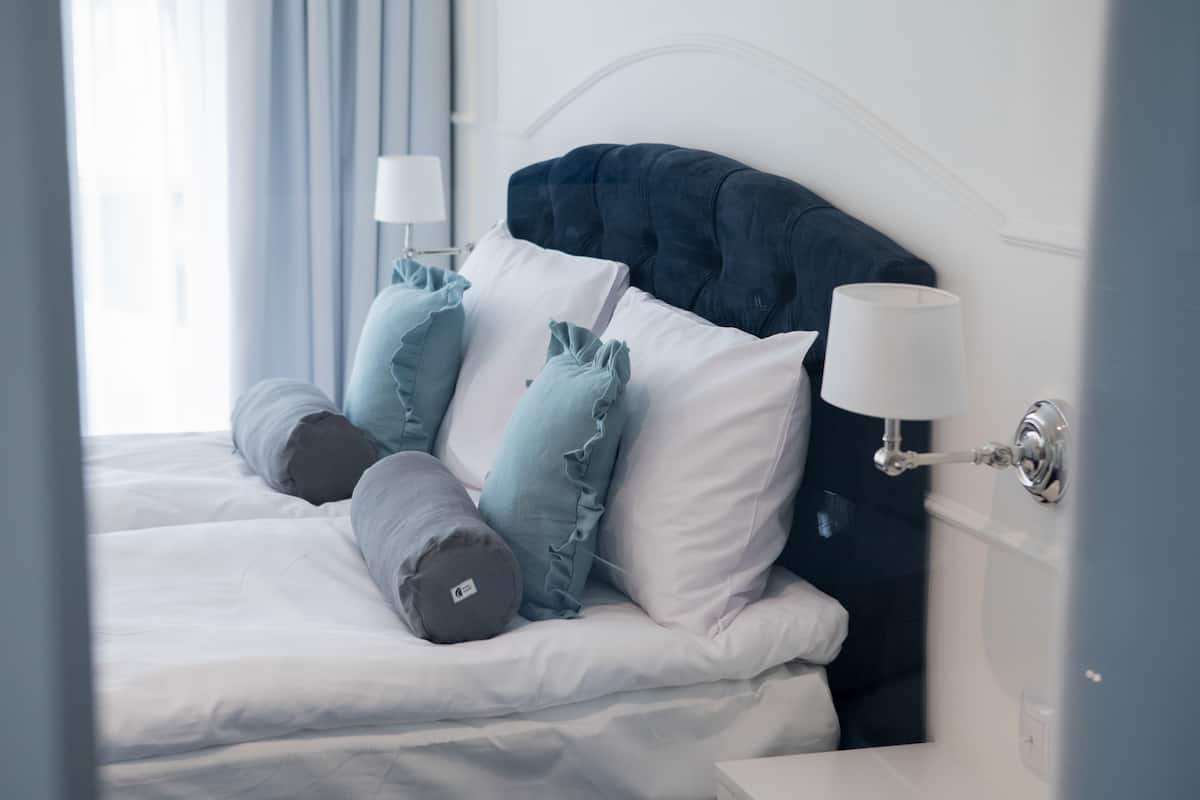 A neatly made bed with white linens and multiple pillows, including a grey cylindrical pillow and light blue ruffled pillows.