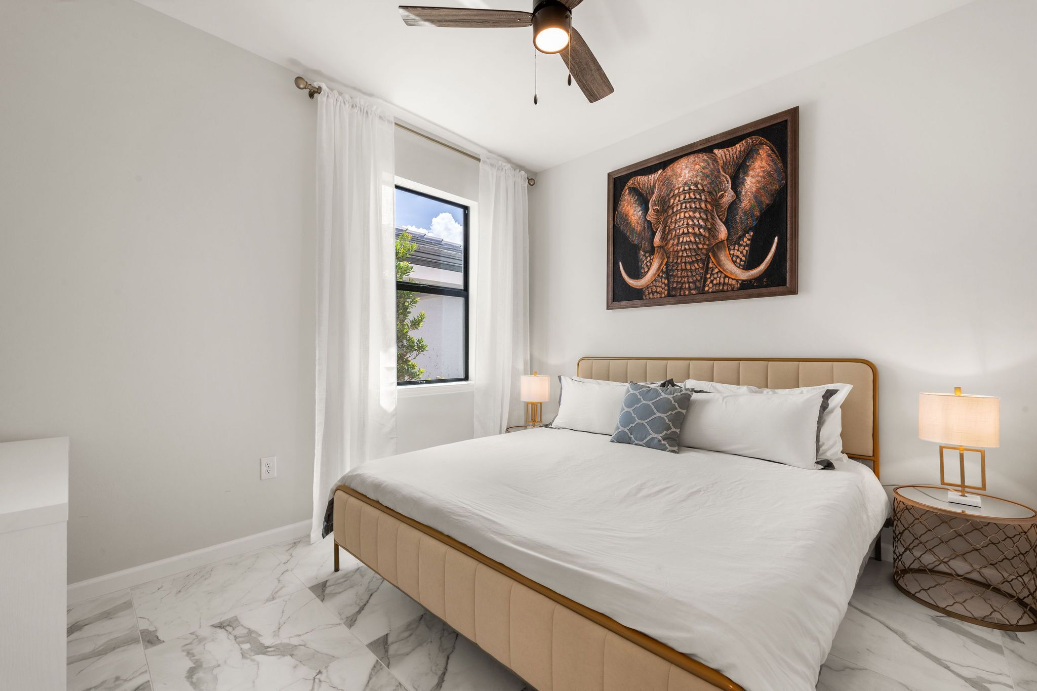 A bedroom with a bed, a ceiling fan, a window with curtains, a wall-mounted elephant artwork, and a bedside lamp.