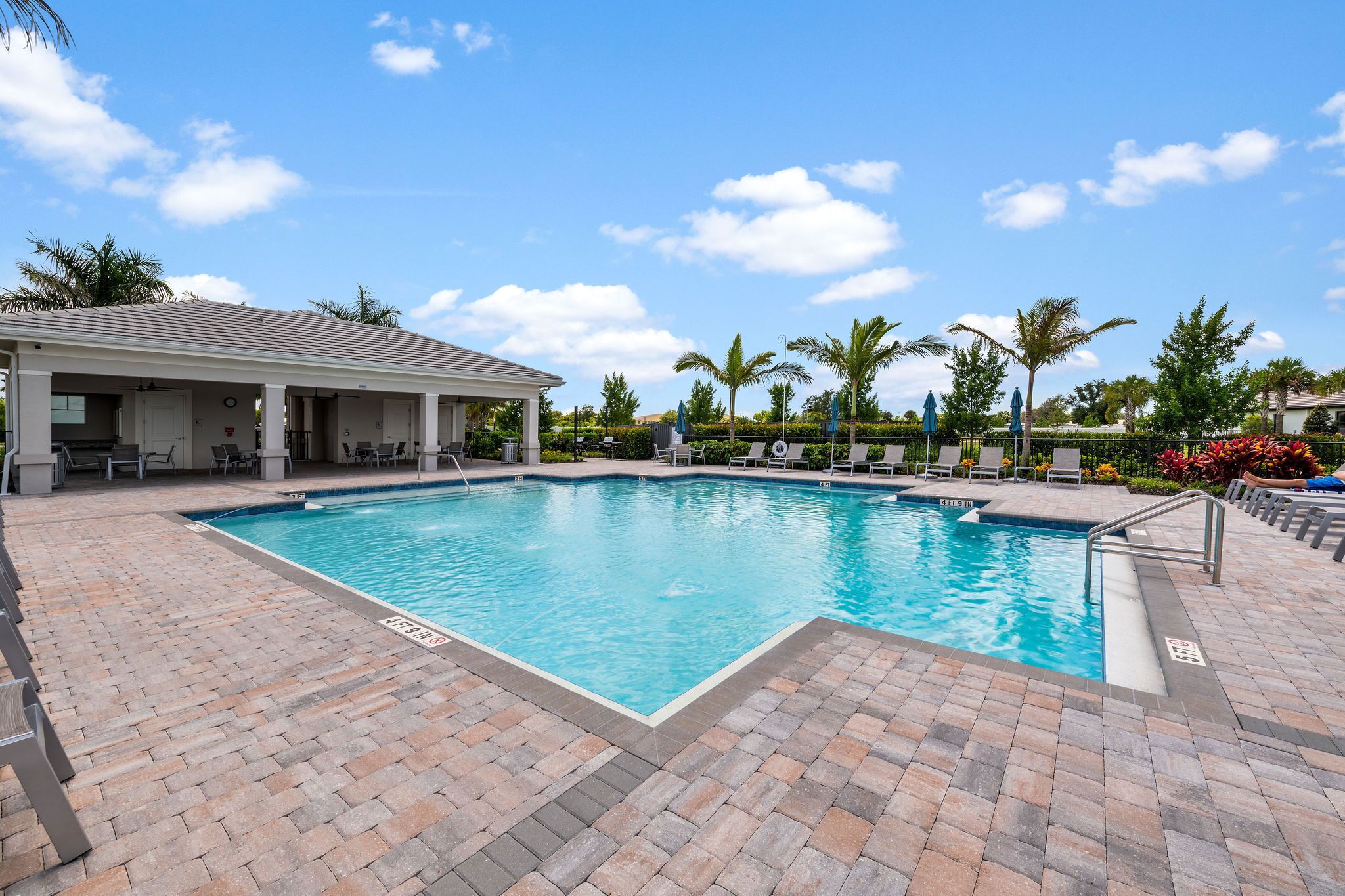 A rectangular swimming pool with a tiled deck, lounge chairs, and a covered seating area.