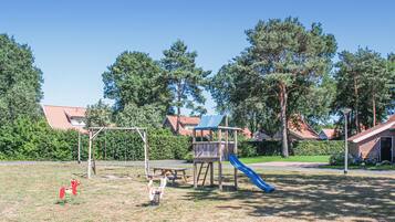 A playground with swings, a slide, and picnic tables in a park with houses and trees in the background.