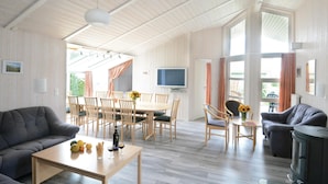 A spacious living area with a wooden ceiling, a dining table with chairs, a sofa, a TV mounted on the wall, and a fireplace. - Strandblick 1 (Brodersby)