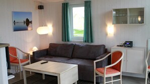 Een kamer met een grijze bank, twee witte stoelen, een klein wit tafeltje, een televisie, een kast met glazen deuren en een raam met gordijnen.