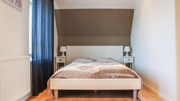 Une chambre à coucher avec un lit, deux tables de chevet, une fenêtre avec des rideaux et un radiateur.