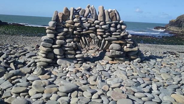 Una escultura de piedra en una playa de guijarros, con el océano y acantilados al fondo.