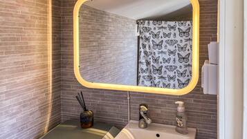 A bathroom with a mirror, a sink, a shower curtain with a butterfly pattern, a vase with a reed diffuser, and a soap dispenser.