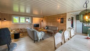 L’intérieur chaleureux d’un chalet avec un plancher en bois, un canapé, une télévision, une table à manger avec des chaises et un coin cuisine équipé d’un réfrigérateur.
