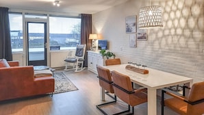 Un salon moderne avec un canapé, une table à manger et des chaises, un téléviseur et une lampe. Une grande fenêtre, ornée de rideaux, offre une vue sur les bâtiments environnants.