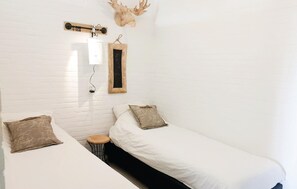 Een kleine kamer met witte muren, twee eenpersoonsbedden, een klein krukje, een houten plank met een hertenkop, en een schoolbord waarop 'ZAN' staat.