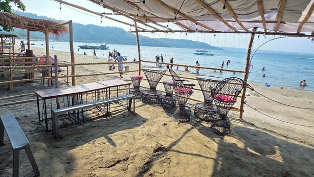 Un espace aménagé avec des tables et des chaises, situé au bord de la plage et abrité par un toit de chaume, d’où l’on a une vue sur la plage et la mer.
