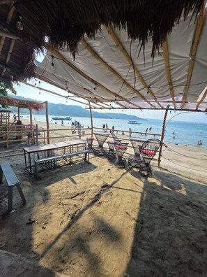 A beachside seating area with tables and chairs under a thatched roof, overlooking the beach and sea. - Wombats Beach Resort (Olongapo)