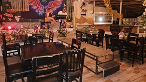 Un restaurant avec des tables et des chaises en bois, une fresque représentant des ailes et des fleurs, et des guirlandes lumineuses suspendues au plafond.