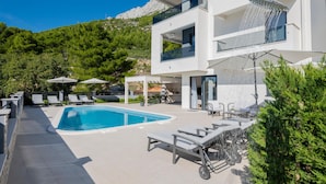 A modern two-story building with a pool, outdoor seating, and a mountain backdrop. - Amazing Apartment in Baska Voda With Wifi, Outdoor Swimming Pool and Heated Swimming Pool (Baska Voda)