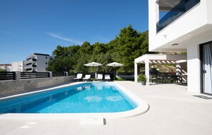 Un área de piscina moderna con piscina de color azul cristalino, sillones blancos y sombrillas.