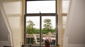Een kamer met een groot raam, een rieten stoel, een rode vaas met bloemen en een tafeltje met een kaars.