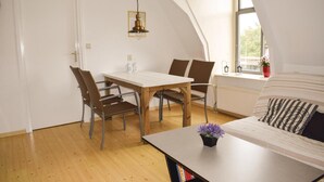 Een gezellige, goed verlichte kamer met een houten eettafel en stoelen, een witte bank en een kleine potplant.