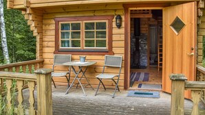 A wooden cabin with a porch, two wicker chairs, a small table, and a window with a view of the surrounding forest.