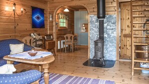 A cozy cabin interior with a wooden floor, a dining area with a table and chairs, a fireplace, and a blue patterned sofa.