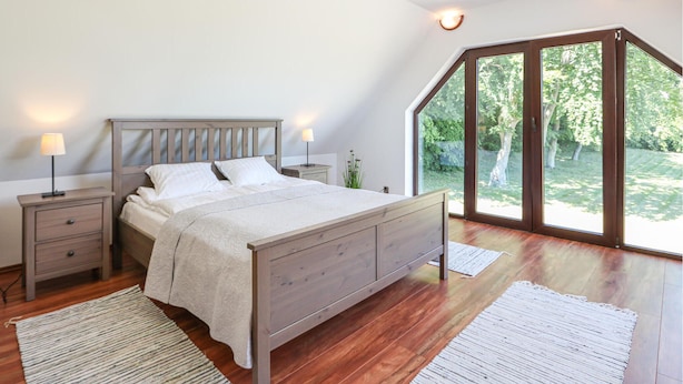 Een slaapkamer met een houten bed, nachtkastjes, een lamp en uitzicht op een tuin door grote ramen.