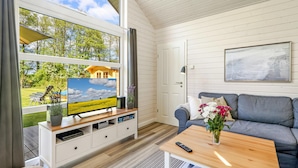 A room with a large window, a television on a wooden stand, a grey sofa, a small table with a vase of flowers, and a door with a framed picture on the wall. - Stunning Home in Krems Ii/warderbrück With 3 Bedrooms and Sauna (Krems II)