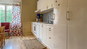 Una cocina compacta con armarios blancos, microondas y lavamanos. Hay una silla de madera y una cortina estampada cerca de la ventana.