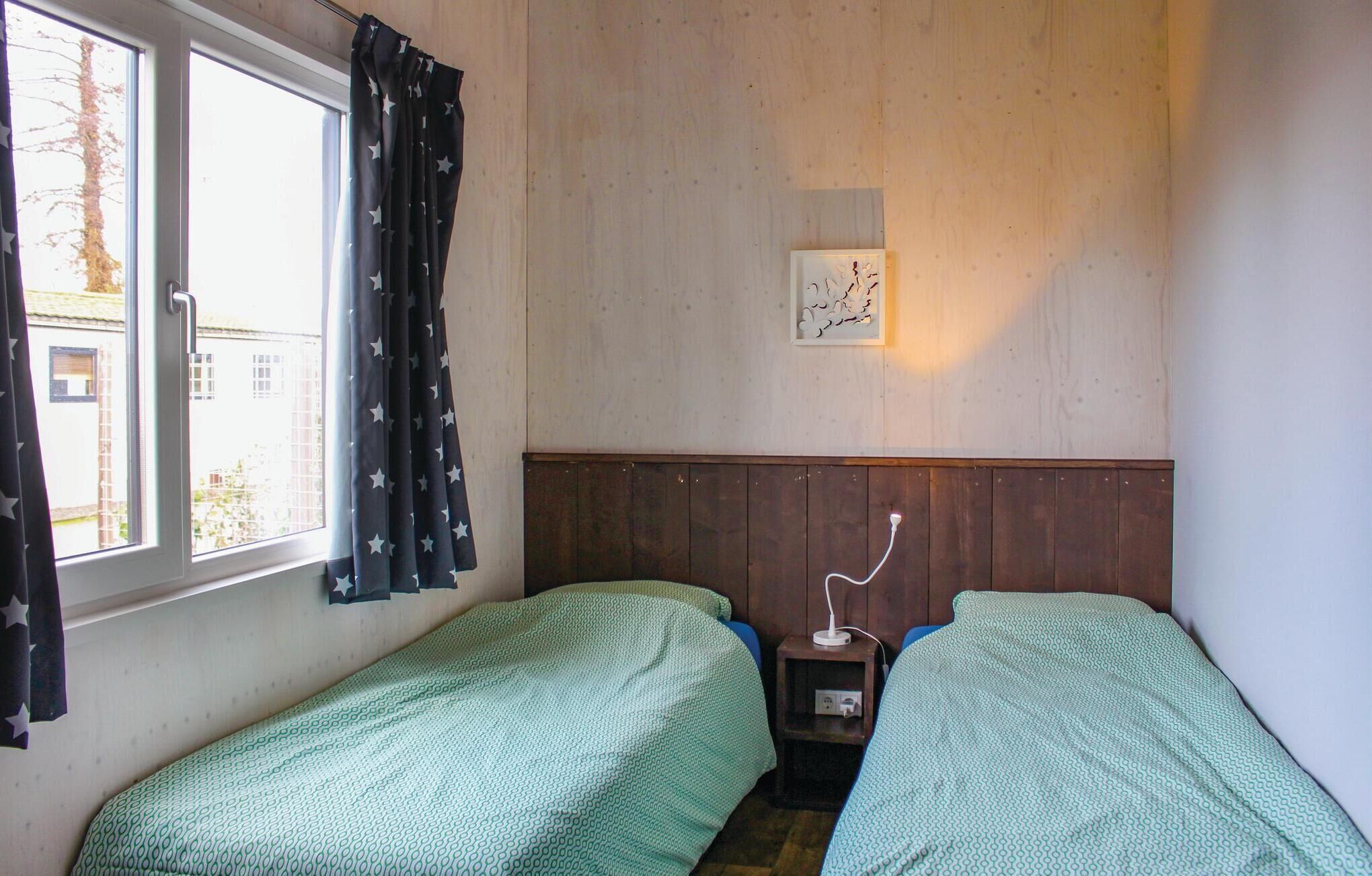 A small, simple room with two single beds, a nightstand with a lamp, a window with curtains, and a wall-mounted picture.