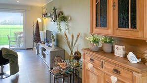 Een woonkamer met een glazen salontafel, een bank en een houten kast met glazen deuren. Er staat een televisie op een dressoir en een plant op de vensterbank.