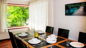 A dining area with a table set for four, a view of greenery through a window, and a painting of a wooden bridge over water on the wall. - Chalet 46 (Hattemerbroek)