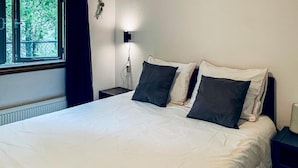 A neatly made bed with white bedding and dark pillows, a bedside lamp, and a window with curtains. - Chalet 46 (Hattemerbroek)
