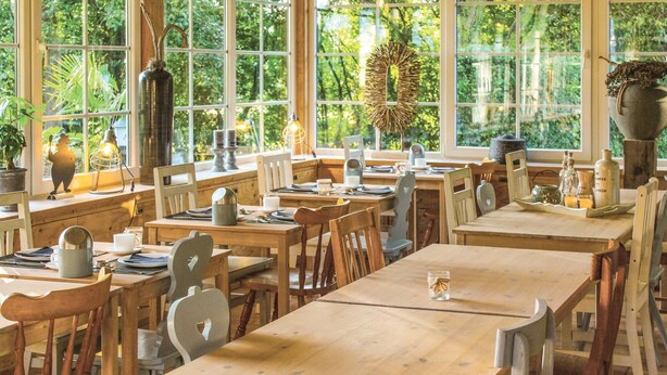 A spacious dining area with wooden tables and chairs, large windows with a view of greenery, and various decorative items on the tables. - Vakantiewoning 11 (Simpelveld)