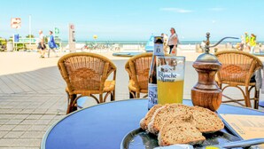 Un tavolo con una bottiglia di Blanquette de Naumer, un bicchiere di succo e fette di pane su un tavolo di un caffè sulla spiaggia, baciato dal sole.