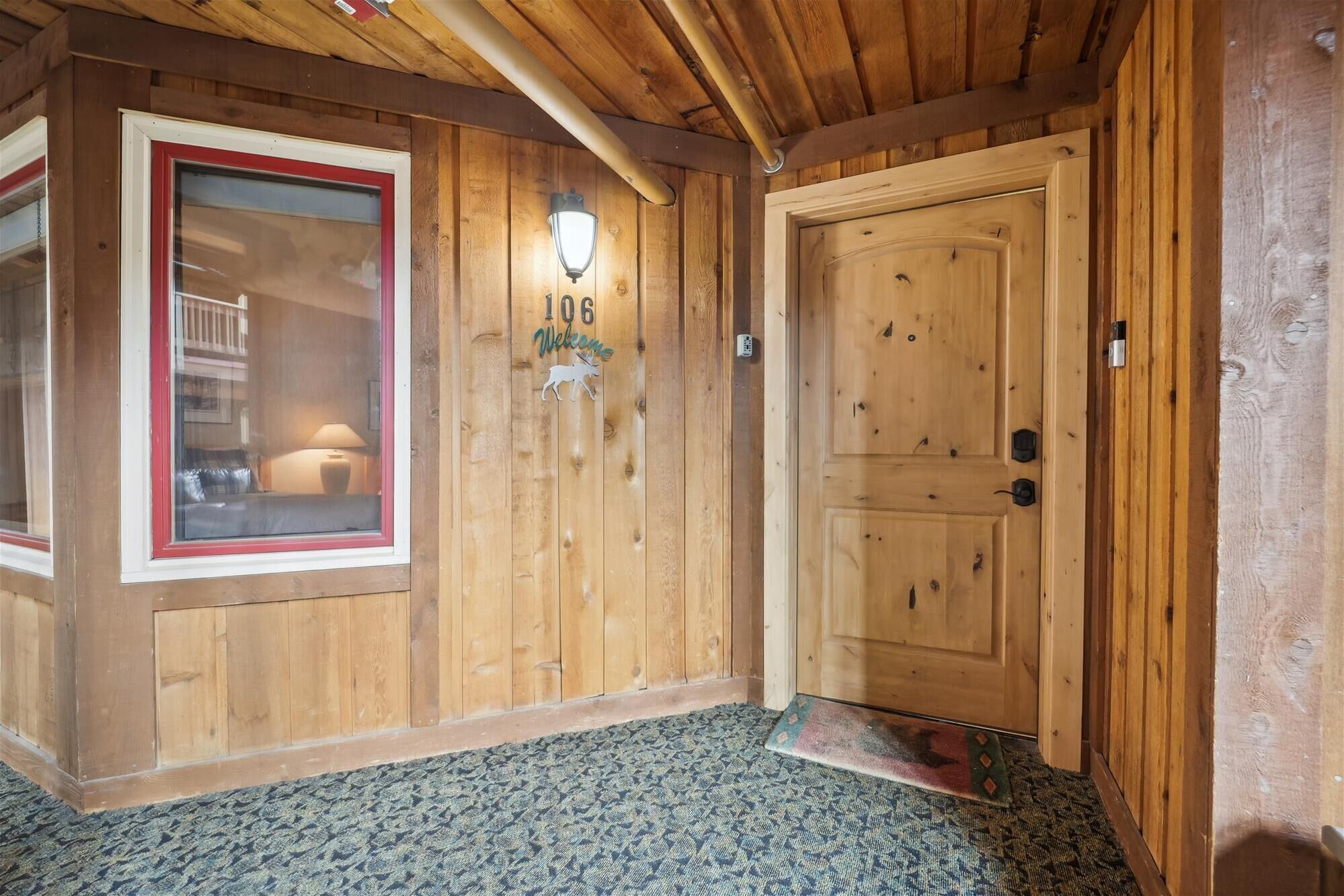 Interior de madeira com uma porta, uma janela com moldura vermelha e uma placa de boas-vindas.