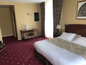 Une chambre d’hôtel avec un grand lit, un bureau avec une chaise, une petite fenêtre et une lampe.